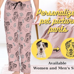 Op de afbeelding: Roze pyjamabroek met een herhalend patroon van een hondenkop, poot afdrukken en botvormen. De hondenkop is zwart-wit. De tekst "Gepersonaliseerde huisdierfoto broek" staat in de rechterbovenhoek van de afbeelding. De tekst "Beschikbaar in dames- en herenmaten" staat in de rechterbenedenhoek van de afbeelding.
