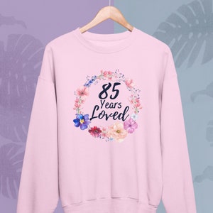 Puede incluir: Sudadera rosa con un diseño de corona floral y el texto "85 Years Loved" en negro.
