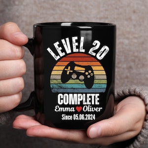 May include: Black coffee mug with a retro-style design featuring a video game controller and the text "Level 20 Complete Emma Oliver Since 05.06.2024".