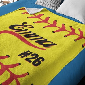May include: Yellow fleece blanket with red stitching and the name "Emma" and number "#26" in black, depicting a softball.