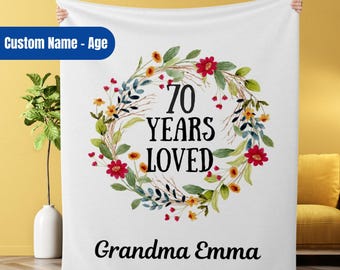 May include: White throw blanket with a floral wreath design. The wreath has green leaves, red, yellow, and pink flowers. The text "70 Years Loved" is in the center of the wreath. The text "Grandma Emma" is at the bottom of the blanket.