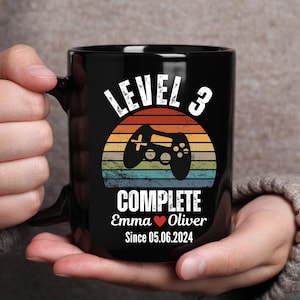 May include: Black coffee mug with a retro-style design featuring a video game controller and the text "Level 3 Complete Emma Oliver Since 05.06.2024".
