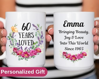 May include: Two white ceramic mugs. One mug has the text "60 YEARS LOVED" with a floral wreath. The other mug says "Emma Bringing Beauty, Joy & Love Into This World Since 1965" with floral accents. The mugs are held by a person.