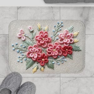 May include: A rectangular beige bath mat featuring an embroidered floral pattern. The flowers are in shades of pink and red, with green leaves and blue details. The mat is on a tiled floor.