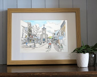 May include: A framed watercolor painting of a town square with a clock tower in the center. The painting is in a light wood frame and is on a wooden shelf. There is a potted plant in the foreground.