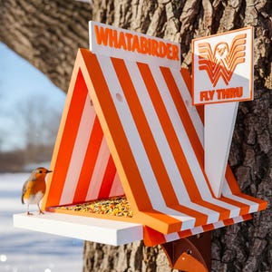 May include: A triangular bird feeder with orange and white stripes, featuring the text WHATABIRDER and a sign reading FLY THRU with a stylized W logo. A small robin stands on the white platform filled with birdseed, attached to a tree trunk.