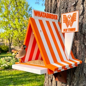 May include: A triangular bird feeder with orange and white vertical stripes, featuring a white base and a sign that reads WHATABIRDER and FLY THRU with a bird logo. A small brown bird stands on the white platform next to a pile of birdseed.