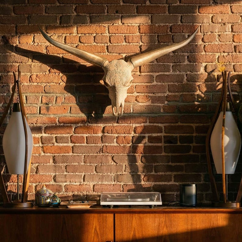 May include: A wall-mounted longhorn steer skull featuring intricate carved patterns across the bone surface. The skull has two long, curved horns extending outwards and is positioned centrally above a wooden console table with lamps.