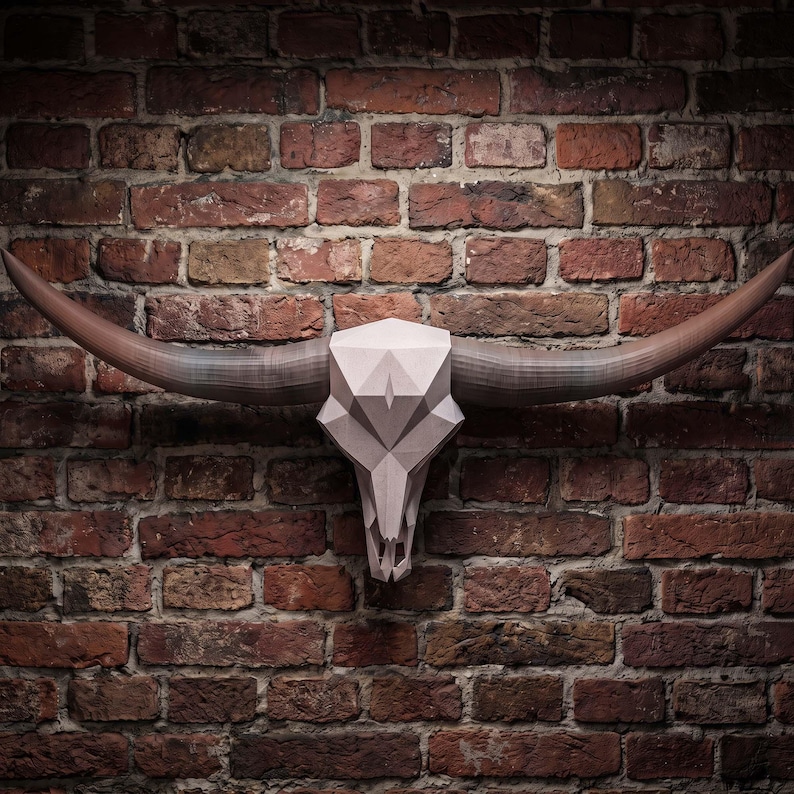 May include: A geometric wall sculpture of a longhorn bull skull featuring a light beige, faceted, low-poly design. The skull has two long, curved, dark brown horns extending horizontally from the sides, creating a wide, symmetrical silhouette.