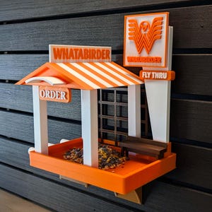 May include: A 3D-printed bird feeder shaped like a fast-food restaurant with an orange and white striped roof. It features the text WHATABIRDER, ORDER, and FLY THRU, a small brown bench, a grid-like back wall, and a tray filled with birdseed.