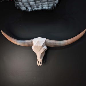 May include: A geometric bull skull wall mount featuring a light beige, speckled, low-poly cranium with long, smooth, curved horns that transition from a light tan at the base to a darker brown at the pointed tips, creating a sharp, angular aesthetic.