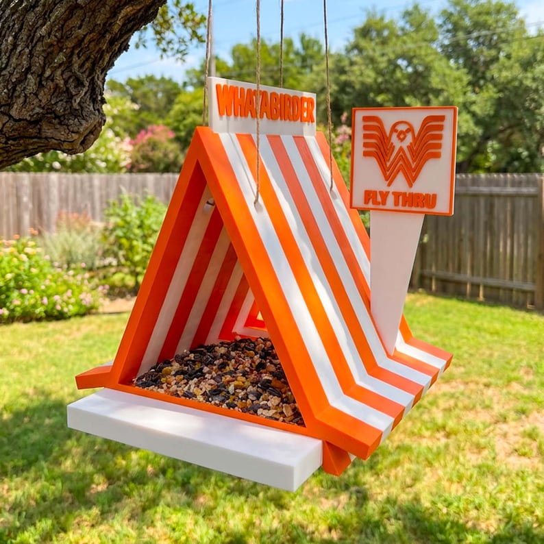 May include: A triangular bird feeder with orange and white stripes, featuring the text "WHATABIRDER" on the roof peak and a square sign attached to the side displaying a stylized "W" logo with the text "FLY THRU" above a tray of birdseed.