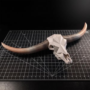 May include: A geometric bull skull sculpture featuring a faceted, light gray cranium and two long, curved horns that transition from a light tan base to a dark gray tip, displayed on a black cutting mat with white grid lines and markings.
