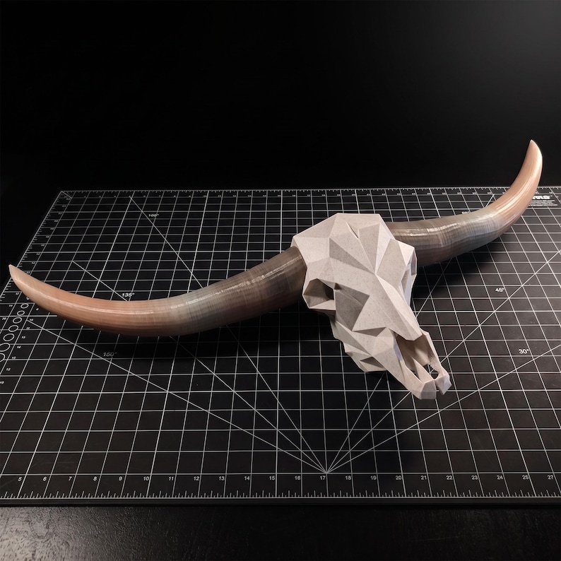 May include: A geometric bull skull sculpture featuring a faceted, light gray cranium and two long, curved horns that transition from a light tan base to a dark gray tip, displayed on a black cutting mat with white grid lines and markings.