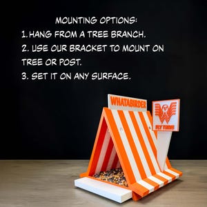 May include: A triangular bird feeder with orange and white stripes, featuring the text WHATABIRDER and a logo with a bird and the text FLY THRU. The text MOUNTING OPTIONS: 1. HANG FROM A TREE BRANCH. 2. USE OUR BRACKET TO MOUNT ON TREE OR POST. 3. SET IT ON ANY SURFACE. is visible.