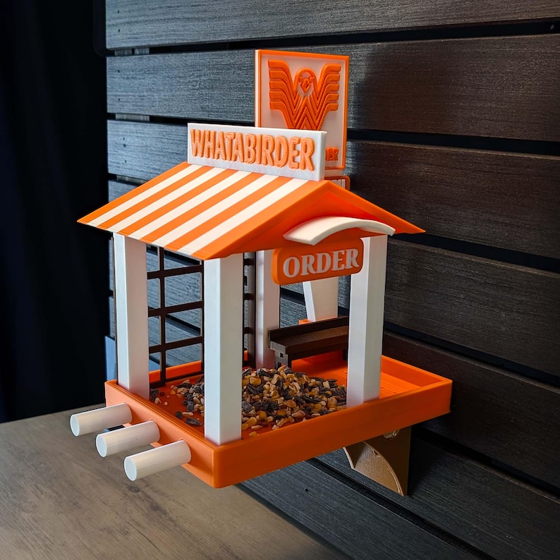 May include: A bird feeder designed like a restaurant with an orange and white striped roof, white pillars, and a sign that reads WHATABIRDER. It features an orange tray filled with birdseed, a small dark bench, and three white cylindrical perches.