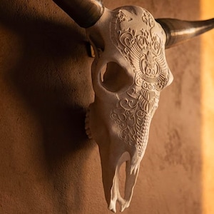 May include: A light-coloured animal skull wall mount featuring intricate, carved floral and geometric patterns across the forehead and snout. The skull has two large, dark, curved horns extending from the sides, creating a striking contrast.