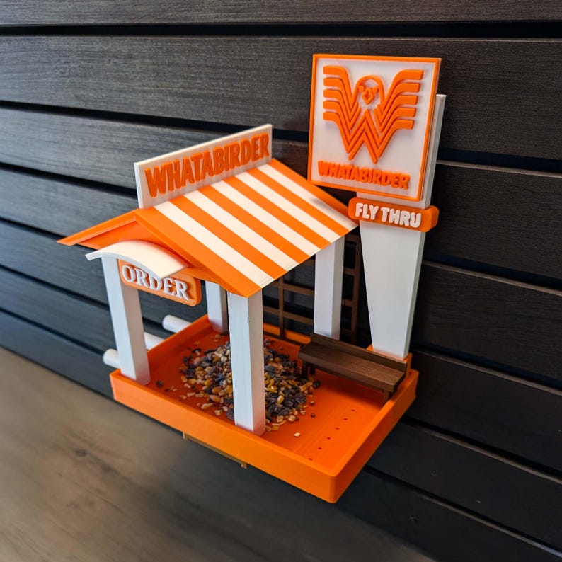 May include: A 3D-printed bird feeder designed as a fast-food restaurant with an orange and white striped roof. It features the text "WHATABIRDER" on the roof and sign, "ORDER" on the side, and "FLY THRU" on the sign, with a small brown bench.