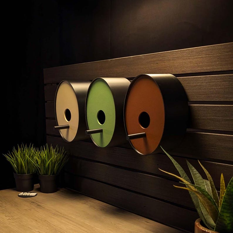 May include: Three cylindrical birdhouses mounted in a horizontal row. Each unit features a black outer shell, a circular entrance hole, and a small perch. The front panels are colored in light beige, sage green, and burnt orange respectively.