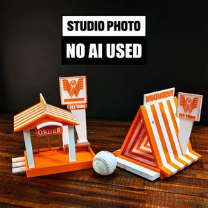 May include: Two orange and white plastic miniature structures featuring the text "WHATABIRDER" and "FLY THRU" with an "ORDER" sign, accompanied by a white baseball. The structures have striped roofs and signage, with one building-style and one A-frame design.