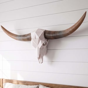 May include: A geometric wall sculpture of a longhorn skull featuring a faceted, light-colored cranium and two long, curved, brown-toned horns that extend horizontally from the sides, creating a sharp, angular, and symmetrical appearance.