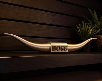 May include: A metallic gold-toned longhorn steer horn sculpture featuring a central rectangular block with the word BOSS embossed in dark lettering. The sculpture has two long, curved, pointed horns extending outward from the center piece.