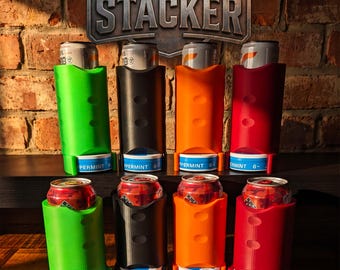 May include: Eight vertical plastic can stackers in green, black, orange, and red, arranged in two rows. Each holder secures a beverage can at the top and a round peppermint tin at the base. A sign above reads THE STACKER in metallic lettering.