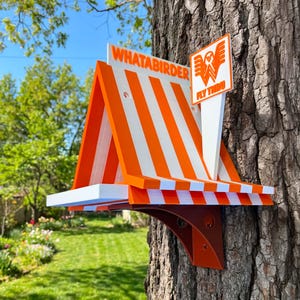 May include: A bird feeder shaped like a restaurant stand with orange and white vertical stripes. It features the text WHATABIRDER on the roof and a square sign with a stylized W logo and the text FLY THRU. The structure is mounted on a tree trunk.