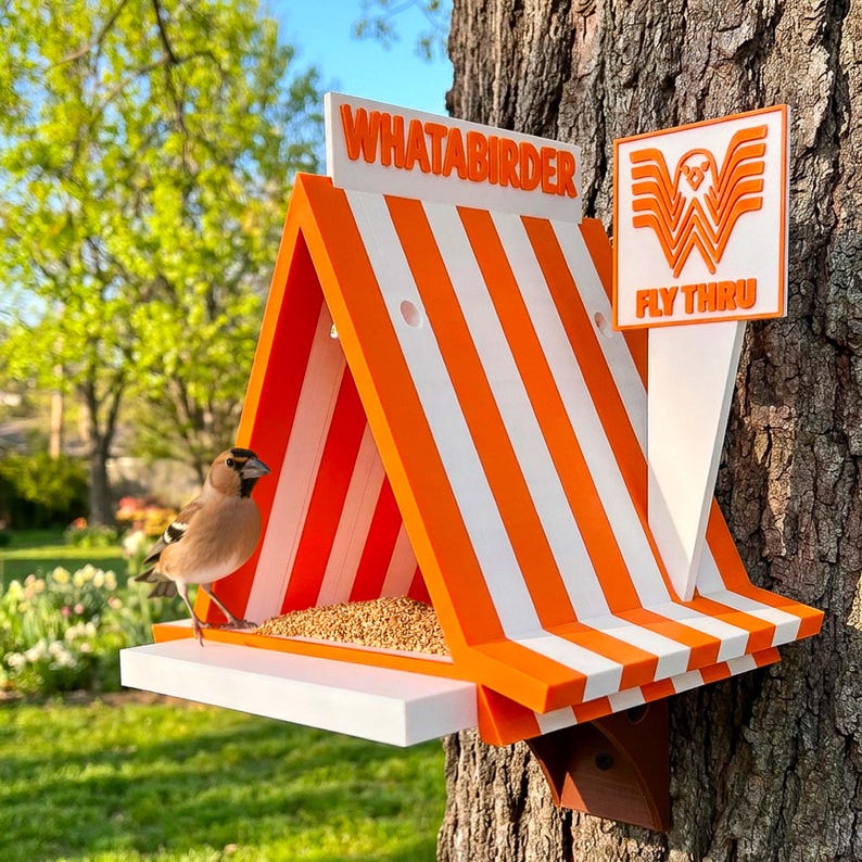 May include: A triangular bird feeder with orange and white vertical stripes, featuring a white base and a sign that reads WHATABIRDER and FLY THRU with a bird logo. A small brown bird stands on the white platform next to a pile of birdseed.