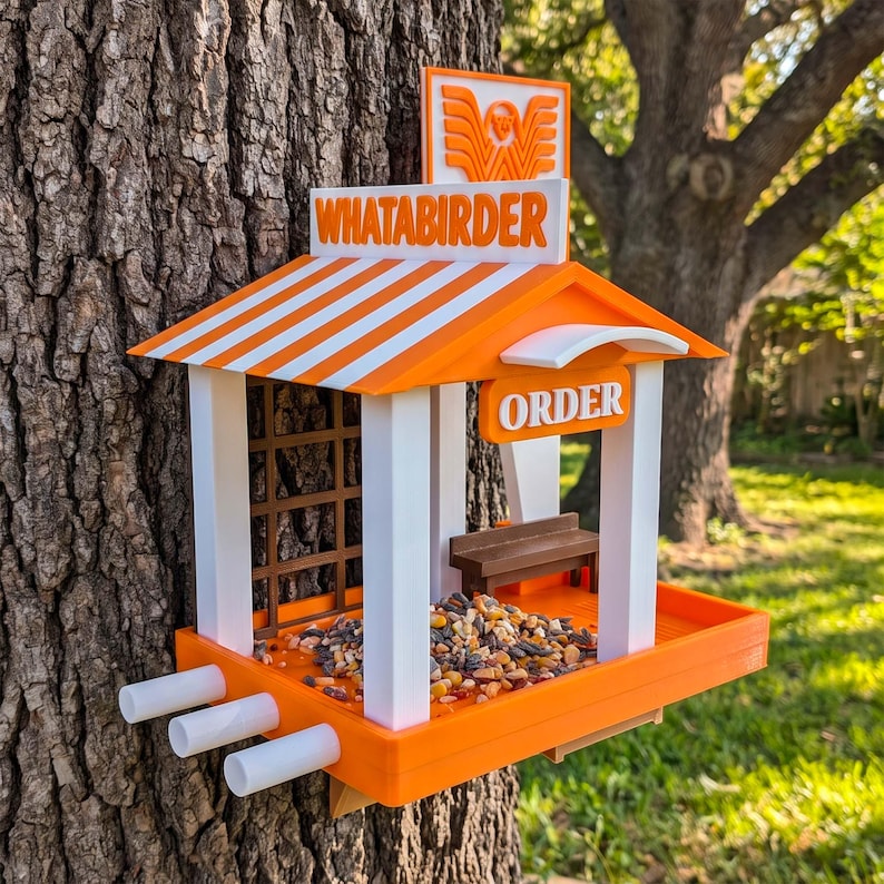 May include: A bird feeder shaped like a fast-food restaurant with an orange and white striped roof, white pillars, and a sign that reads WHATABIRDER. The structure features an ORDER sign, a small brown bench, and a tray filled with birdseed.