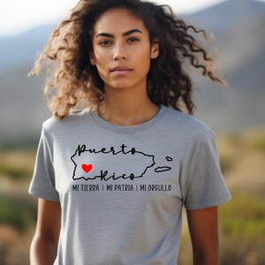 Puede incluir: Una camiseta gris con un contorno negro de Puerto Rico y un coraz&oacute;n rojo en el centro. El texto "Puerto Rico" est&aacute; escrito encima del contorno y el texto "Mi tierra | Mi patria | Mi orgullo" est&aacute; escrito debajo del contorno.