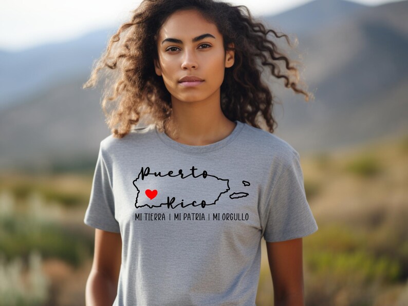 Puede incluir: Una camiseta gris con un contorno negro de Puerto Rico y un coraz&oacute;n rojo en el centro. El texto "Puerto Rico" est&aacute; escrito encima del contorno y el texto "Mi tierra | Mi patria | Mi orgullo" est&aacute; escrito debajo del contorno.