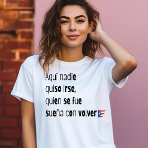 May include: White t-shirt with the text "Aquí nadie quiso irse, quien se fue sueña con volver" and a Puerto Rican flag graphic. The shirt is worn by a person with brown hair, posing in front of a brick wall and a pink wall.
