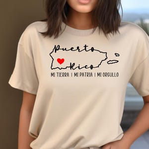 Puede incluir: Una camiseta beige con un contorno negro de Puerto Rico y un coraz&oacute;n rojo en el centro. El texto "Puerto Rico" est&aacute; escrito en cursiva negra sobre el contorno y el texto "Mi tierra, mi patria, mi orgullo" est&aacute; escrito en negro debajo del contorno.