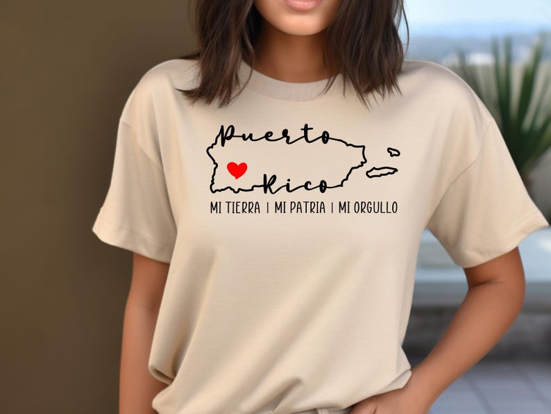 Puede incluir: Una camiseta beige con un contorno negro de Puerto Rico y un coraz&oacute;n rojo en el centro. El texto "Puerto Rico" est&aacute; escrito en cursiva negra sobre el contorno y el texto "Mi tierra, mi patria, mi orgullo" est&aacute; escrito en negro debajo del contorno.