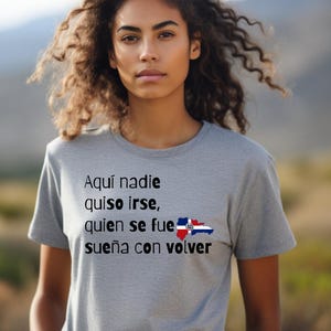 May include: A gray t-shirt with the text "Aquí nadie quiso irse, quien se fue sueña con volver" and a Dominican Republic flag graphic. The shirt is worn by a person with curly hair.
