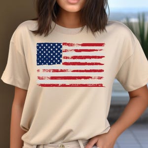 May include: A beige t-shirt featuring a distressed American flag design. The flag has a blue square with white stars in the upper left corner and red and white stripes. The shirt is a casual style.