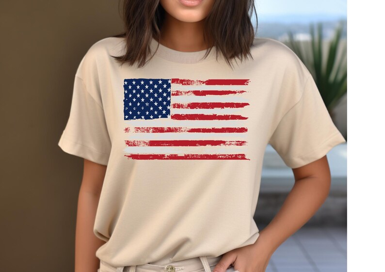 May include: A beige t-shirt featuring a distressed American flag design. The flag has a blue square with white stars in the upper left corner and red and white stripes. The shirt is a casual style.