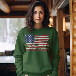May include: A green sweatshirt featuring a distressed American flag design. The flag has a blue field with white stars and red and white stripes. The sweatshirt is worn by a person with dark hair.
