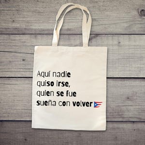 May include: A white tote bag with black text that reads "Aquí nadie quiso irse, quien se fue sueña con volver" and a Puerto Rican flag. The bag has two handles and is made of a canvas-like material.