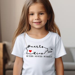 Puede incluir: Una camiseta blanca con un contorno negro de Puerto Rico y las palabras "Puerto Rico" con un coraz&oacute;n rojo en el medio. El texto debajo del contorno dice "Mi tierra, mi patria, mi orgullo."