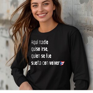 May include: Black long-sleeve shirt with white text that reads "Aquí nadie quiso irse, quien se fue sueña con volver" and a Puerto Rican flag. The shirt is worn by a person with long brown hair.