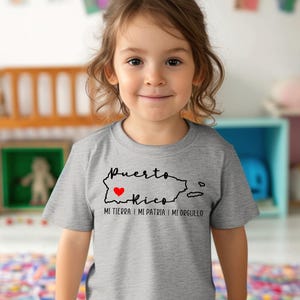 Puede incluir: Un ni&ntilde;o o ni&ntilde;a con una camiseta gris con un contorno negro de Puerto Rico y las palabras "Puerto Rico Mi Tierra I Mi Patria I Mi Orgullo" con un coraz&oacute;n rojo sobre la isla.