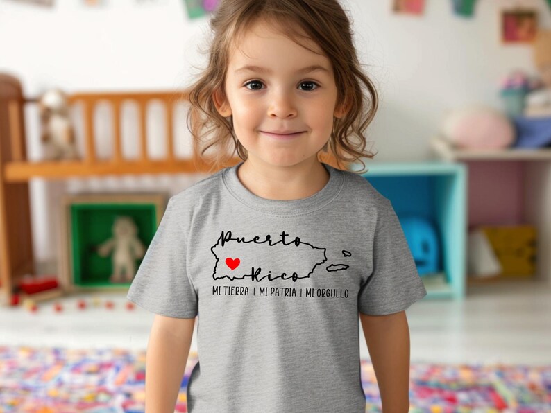 Puede incluir: Un ni&ntilde;o o ni&ntilde;a con una camiseta gris con un contorno negro de Puerto Rico y las palabras "Puerto Rico Mi Tierra I Mi Patria I Mi Orgullo" con un coraz&oacute;n rojo sobre la isla.