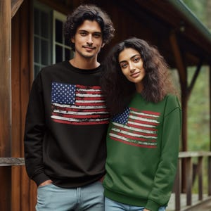 May include: A couple wearing matching sweatshirts with a distressed American flag design. The sweatshirt on the left is black, and the one on the right is green. Both are wearing blue jeans.