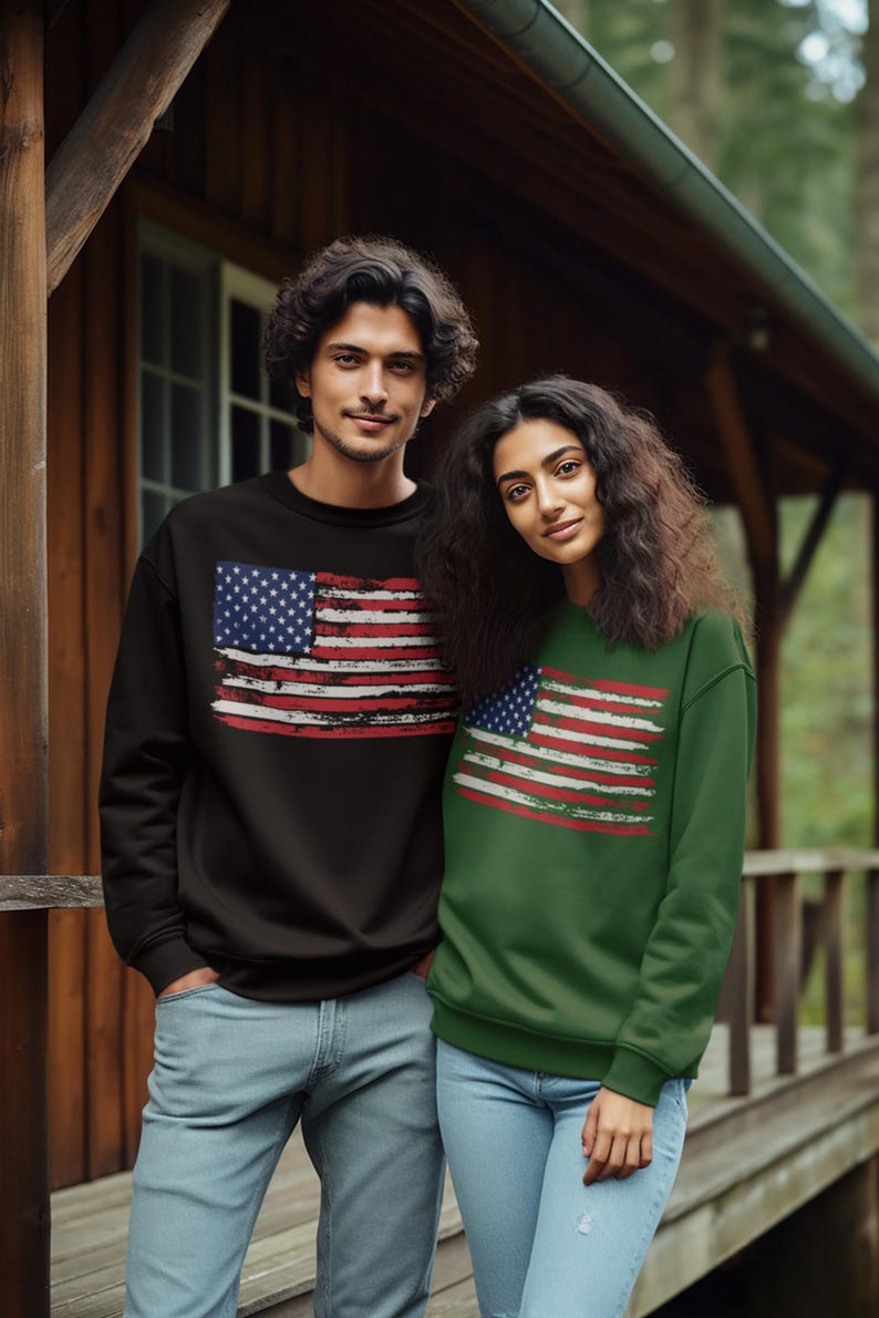 May include: A couple wearing matching sweatshirts with a distressed American flag design. The sweatshirt on the left is black, and the one on the right is green. Both are wearing blue jeans.