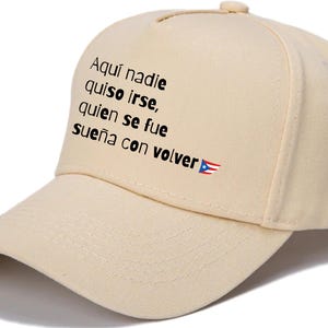 May include: A beige baseball cap with the text "Aquí nadie quiso irse, quien se fue sueña con volver" in black, along with a Puerto Rican flag. The cap has a curved brim and a button on top.