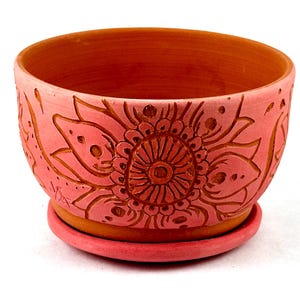 May include: A handcrafted, coral-colored ceramic planter with a matching saucer. The planter features a detailed floral design carved into the exterior. The interior is a darker orange hue, creating a contrast. Ideal for small plants.