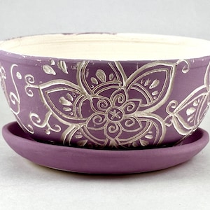 May include: A purple ceramic planter with a matching saucer. The planter features a raised, silver floral design and a white interior. The design includes a large flower and swirling patterns. The planter is suitable for indoor or outdoor use.