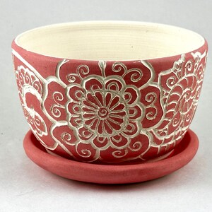 May include: A coral-colored ceramic planter with a matching saucer. The planter features a detailed, raised floral design in white. The interior of the planter is a cream color, creating a contrast with the exterior.